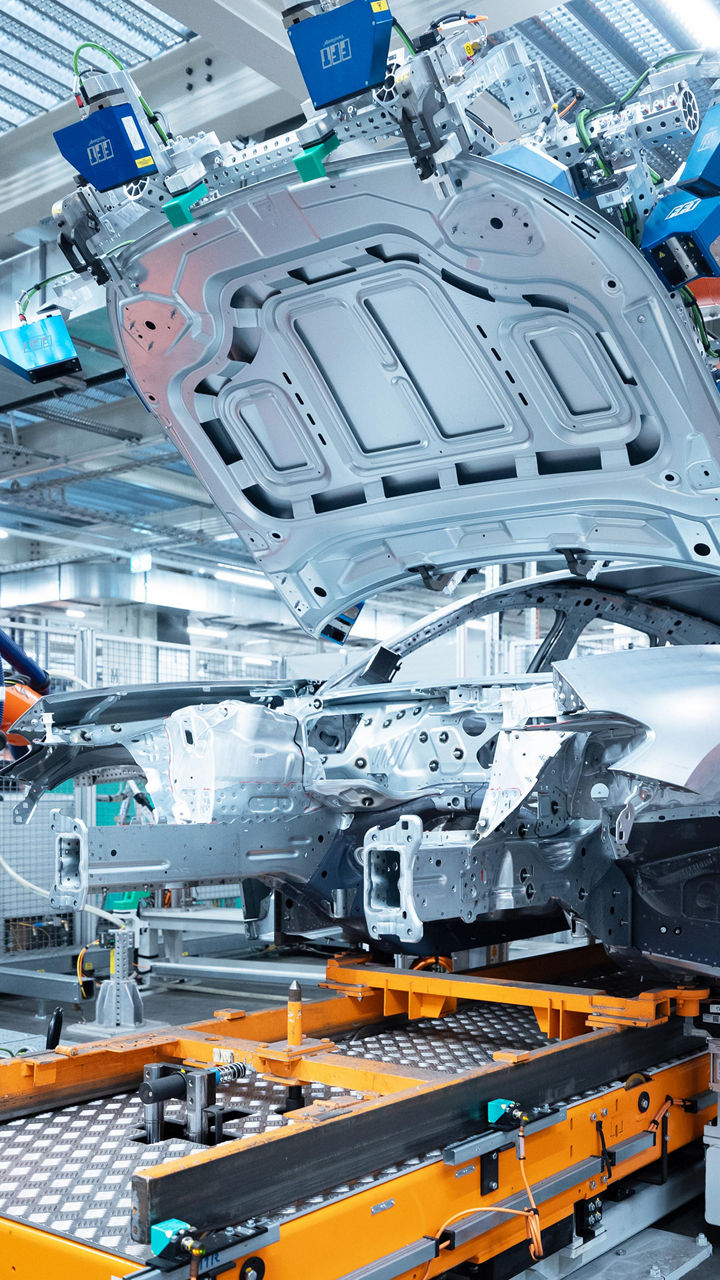 A modern production robot in a production hall lifts the body of a car. Surrounded by industrial machinery and lighting, the image shows a high-tech assembly process.