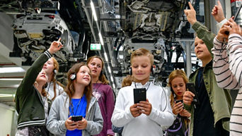 A group of children with headsets are standing under production facilities and looking upwards.