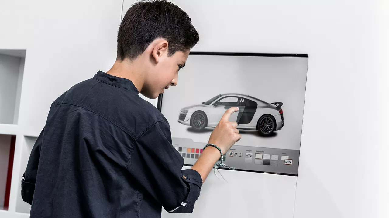 Junge bedient einen Touchscreen mit Auto-Konfigurator, auf dem ein silbernes Sportauto angezeigt wird.