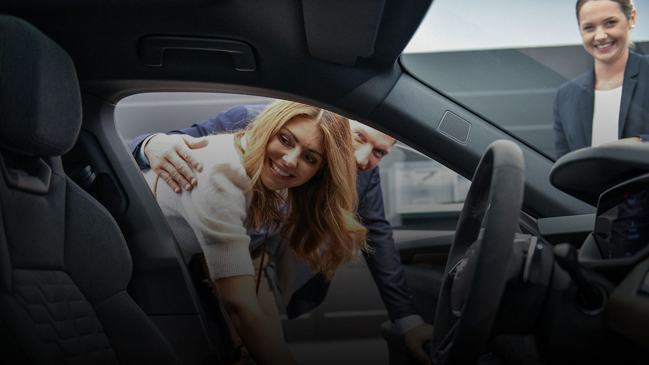 A woman leans into a car with a smile, accompanied by a man and a woman watching her from outside. The scene appears friendly and inviting, in a car dealership or similar environment.