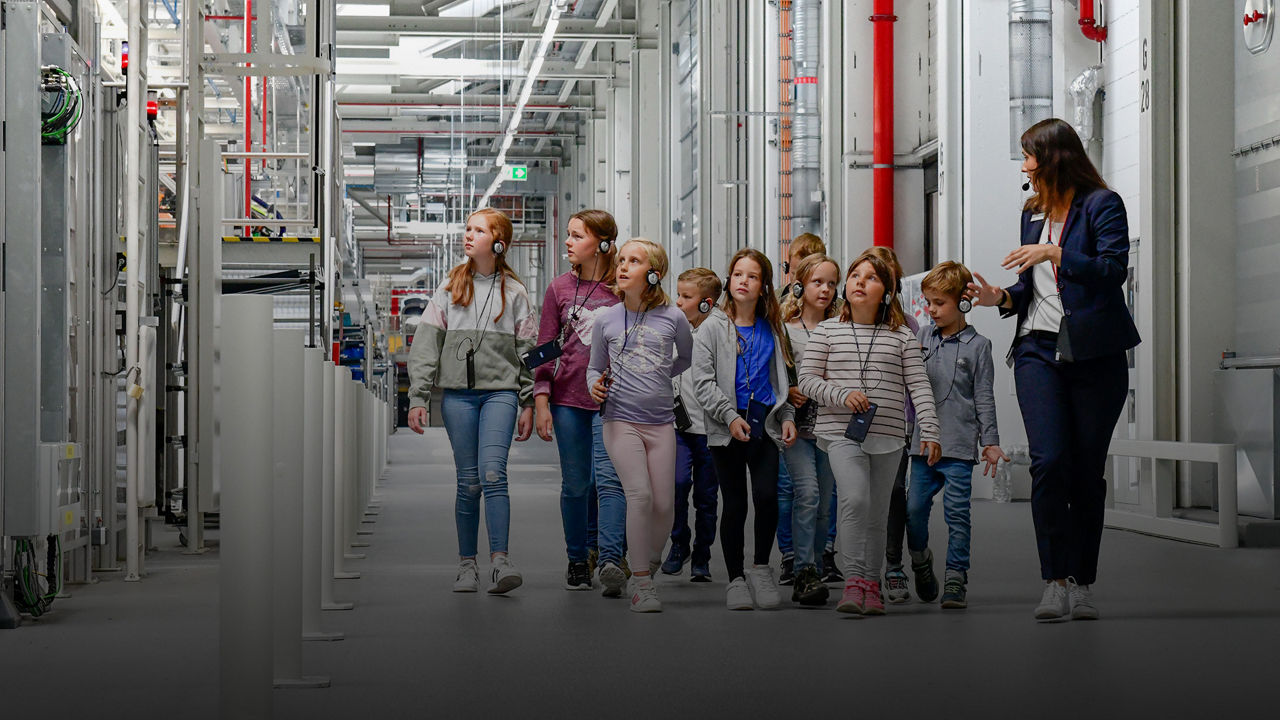 A group of children are standing in a production hall looking at the production line. They are wearing headphones and using devices for a guided tour. The children seem interested and curious.