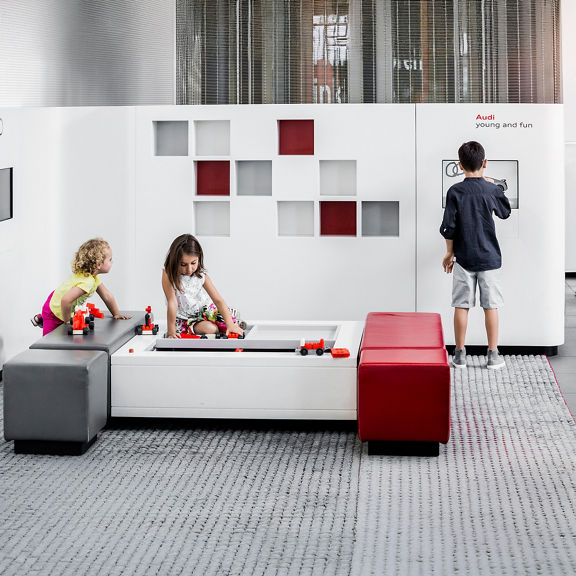 Three children play in a modern play corner with white, red and gray furniture and building blocks.