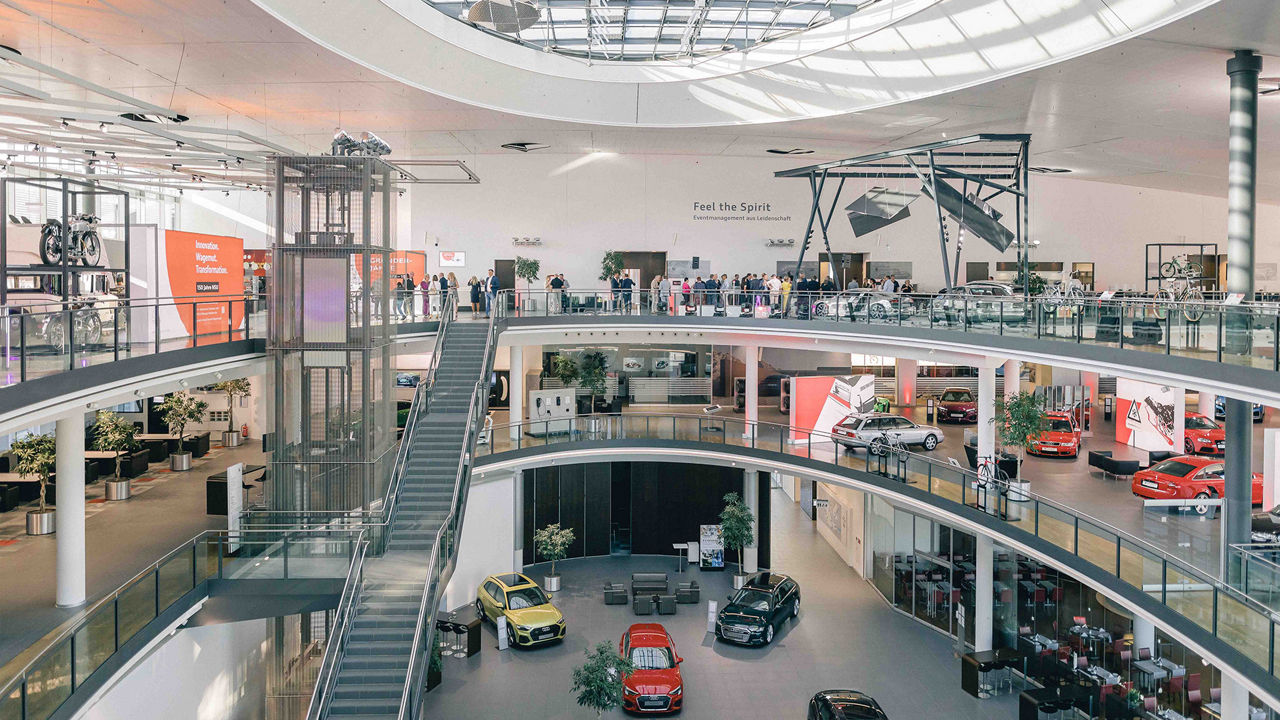 A spacious, modern showroom with several levels and a glass façade. Various Audi vehicles are on display on the ground floor, while the upper floors house offices and exhibition areas. Bright lighting and clear architecture dominate the scene.