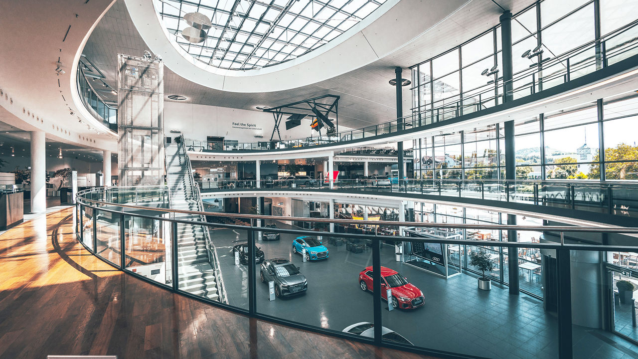 A spacious, modern showroom with several levels and a glass façade. Various Audi vehicles are on display on the ground floor, while the upper floors house offices and exhibition areas. Bright lighting and clear architecture dominate the scene.