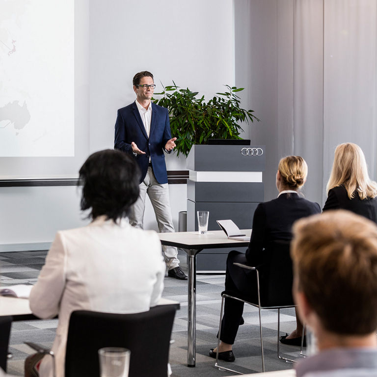 A man is giving a presentation to a group of people in a conference room. A projection screen with a world map and percentages can be seen in the background.