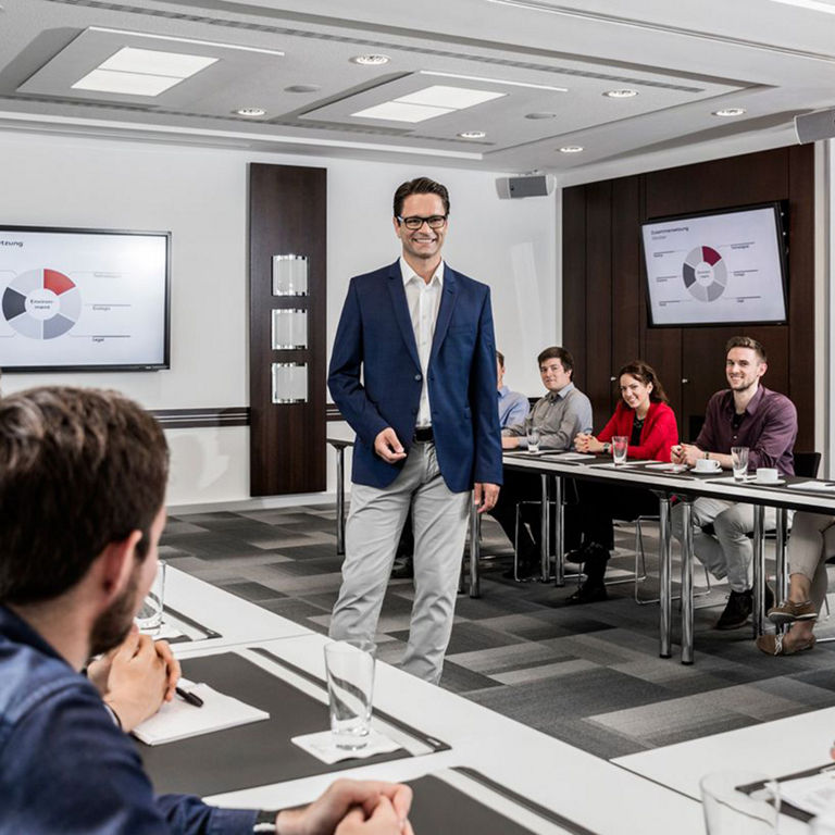 A man in a suit is giving a presentation in a conference room. The participants sit at tables and listen attentively. Diagrams can be seen on screens in the background. The room is furnished in a modern style. 