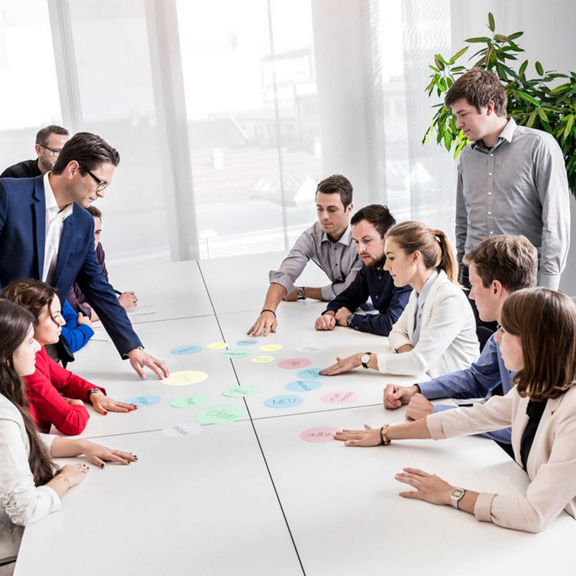 Gruppe sitzt an weißen Tischen in einem modernen Konferenzraum und arbeitet gemeinsam mit bunten Moderationskarten.