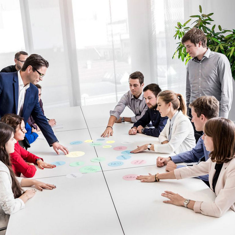 A group of people are sitting and standing around a conference table. They are working together with colourful moderation cards laid out on the table. A man in a suit is moderating the discussion. Plants and large windows can be seen in the background.      