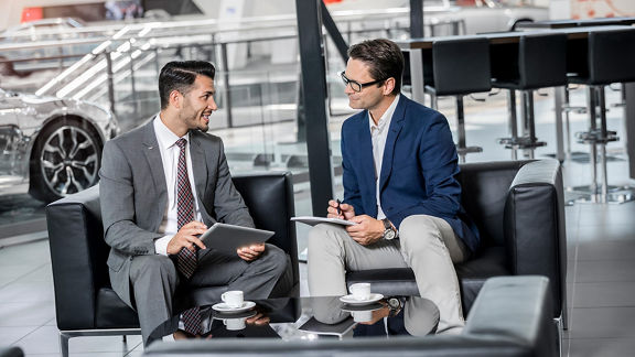 Two men in suits holding a conversation with notes and tablets in a modern lounge with coffee cups, an Audi model in the background.