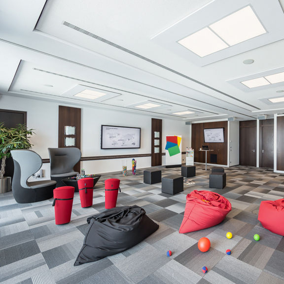 Großer Konferenzraum mit roten und schwarzen Sitzsäcken, Hockern, Displays und bunten Bällen auf dem Boden.