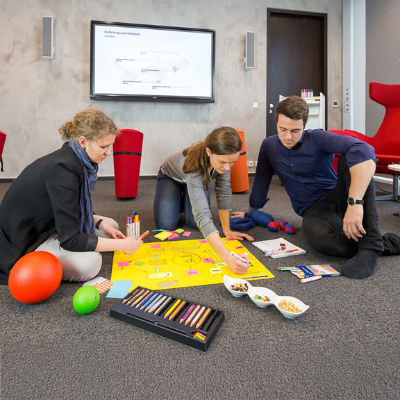 Drei Personen sitzen auf dem Boden in einem Büro, schreiben an Plakat, umgeben von Stiften und Snacks.