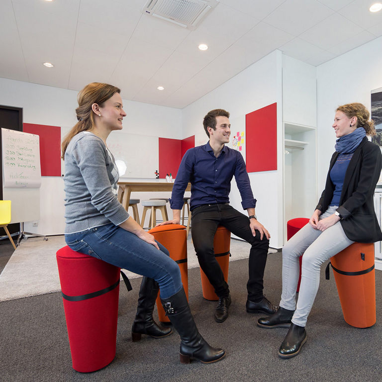 Three people sit on colourful stools in a modern creative space and chat. A high bar, whiteboards and a small kitchenette with drinks can be seen in the background. The room is bright and friendly.