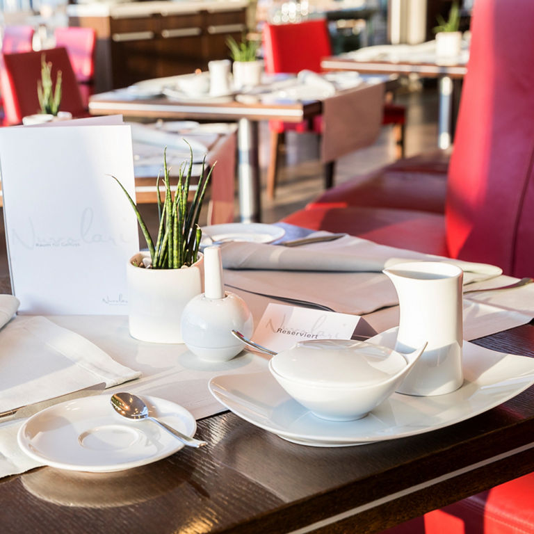 A table set with white porcelain crockery, cloth napkins and a small potted plant as decoration, in a modern restaurant.