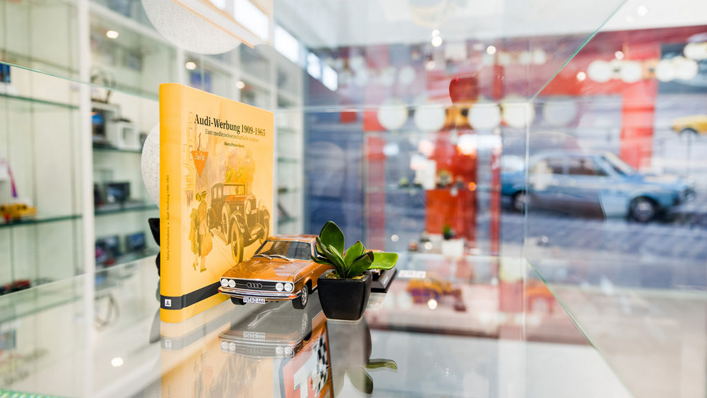 A glass shelf with collector's items, including a model car and a book about Audi advertising, decorated with a small plant.