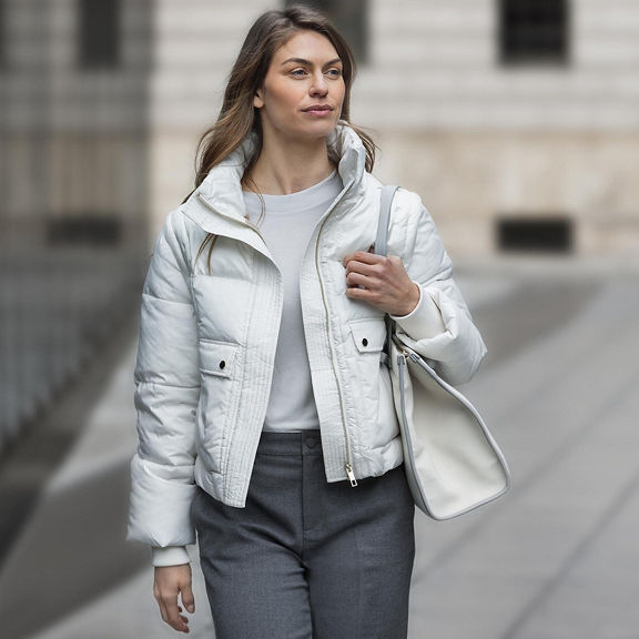 A woman in a white jacket carrying a white leather handbag walks along a footpath next to a mirrored commercial building.