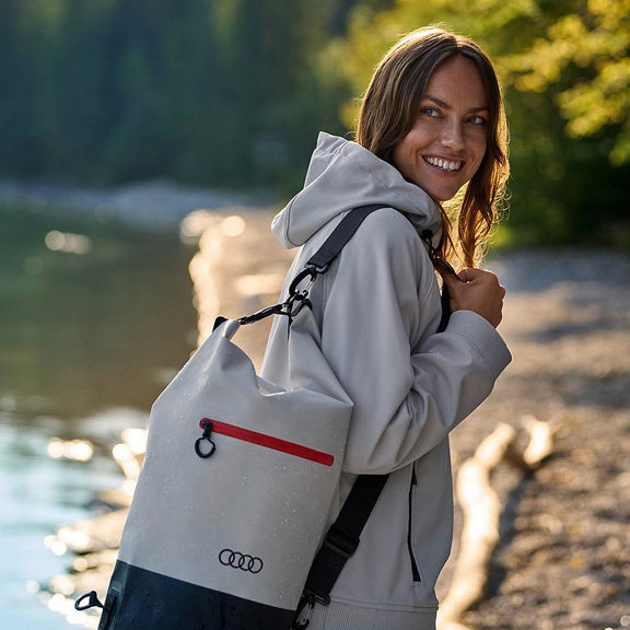 A woman walks along a riverbank and looks into the camera. She is carrying a duffel bag with the Audi emblem around her shoulder.
