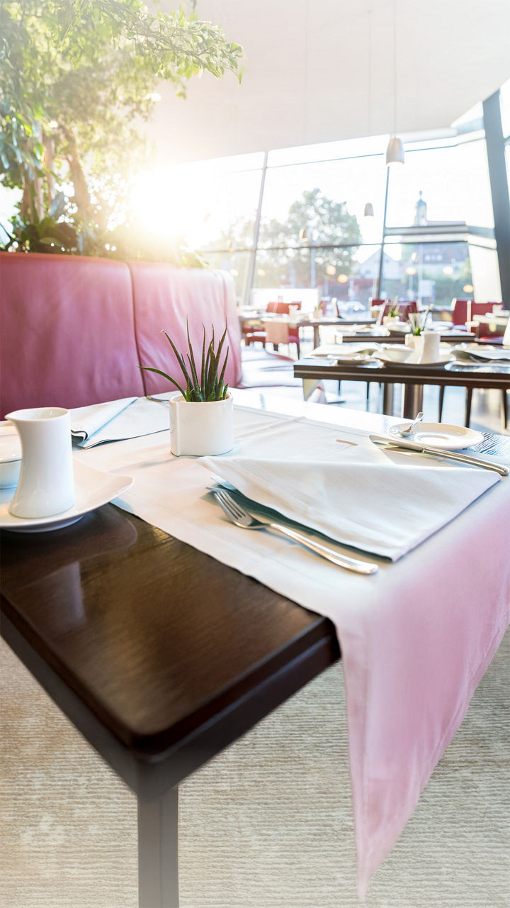 Gedeckter Restauranttisch mit weißem Tischtuch, Geschirr und kleinem Pflanzentopf, im Hintergrund rote Sitzbänke und Sonnenlicht.