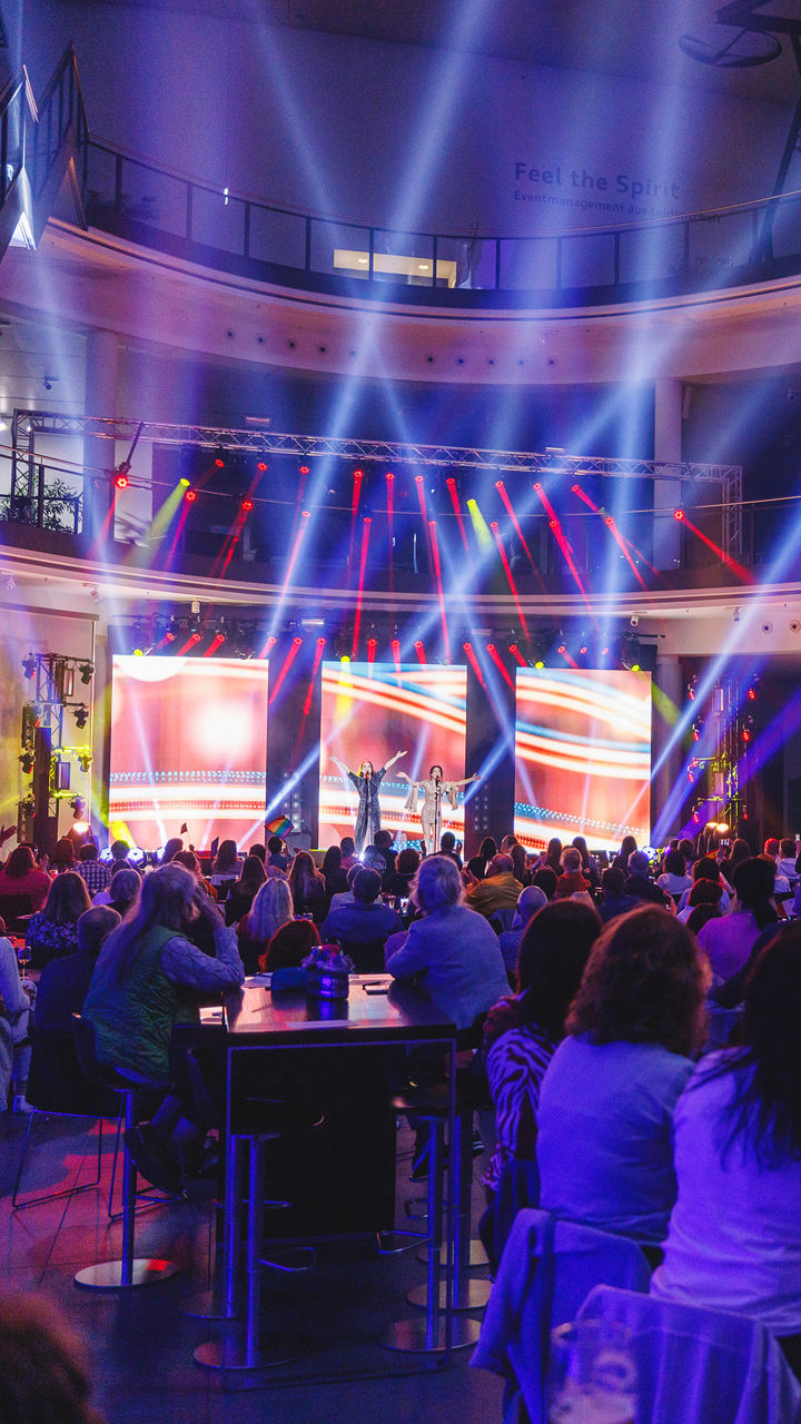 An event in a modern hall with colourful beams of light and large LED screens on the stage. People sit at tables and follow the programme, while the atmosphere is characterised by atmospheric lighting.