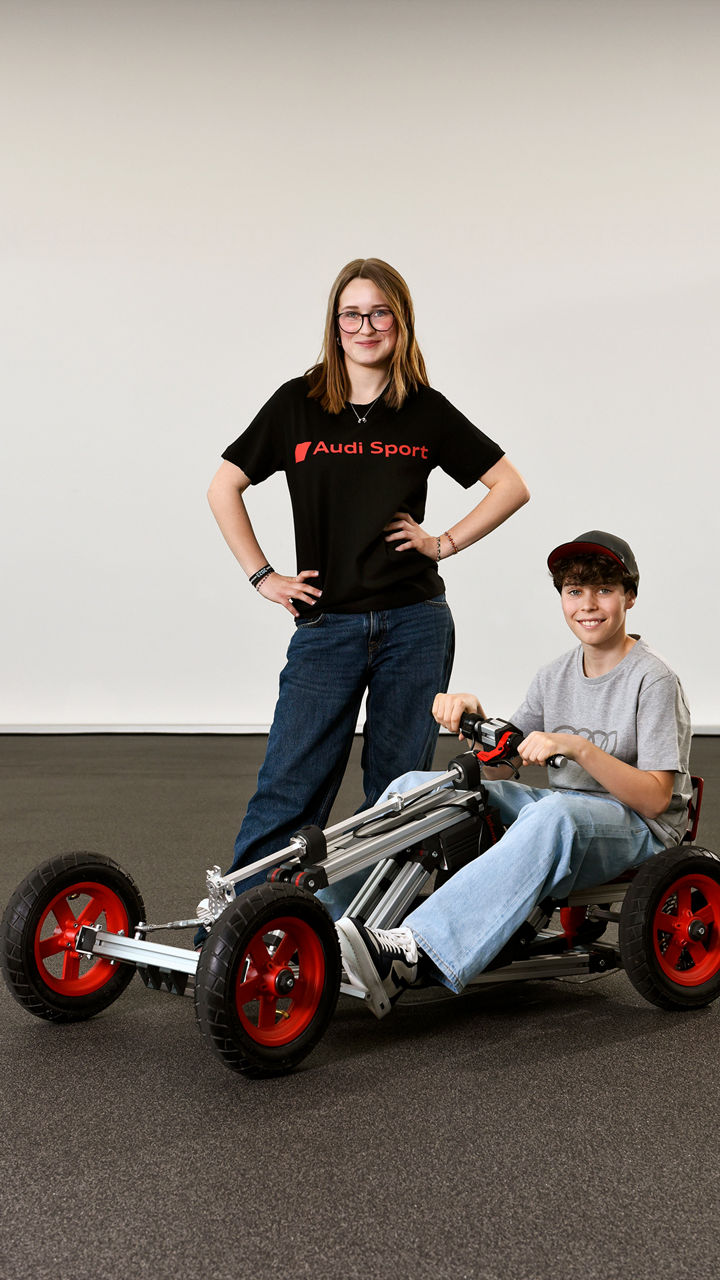 Ein Junge und ein Mädchen montieren gemeinsam ein großes Fahrzeugmodell mit roten Rädern an einem weißen Tisch.