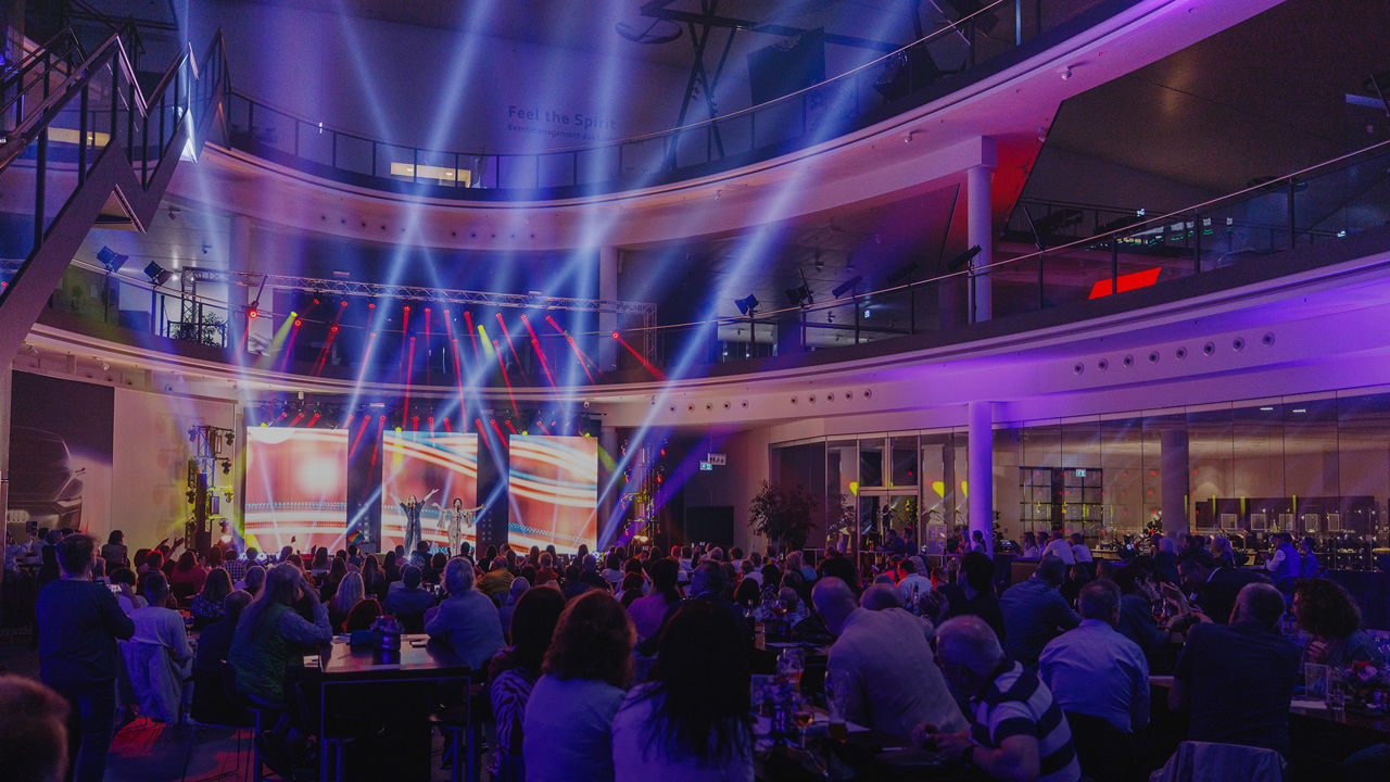 An event in a modern hall with colourful beams of light and large LED screens on the stage. People sit at tables and follow the programme, while the atmosphere is characterised by atmospheric lighting.