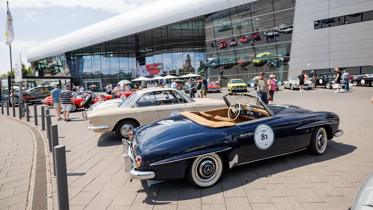Oldtimer-Ausstellung vor modernem Glasgebäude, zahlreiche Menschen betrachten die historischen Fahrzeuge bei Sonnenschein.