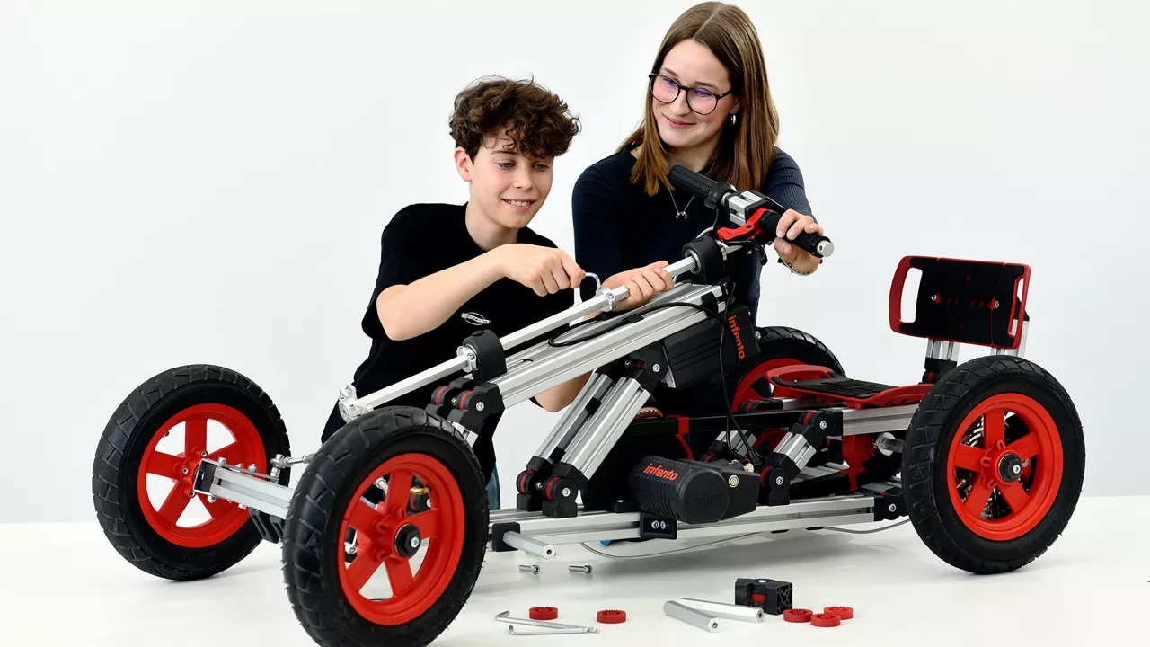 Ein Junge und ein Mädchen montieren gemeinsam ein großes Fahrzeugmodell mit roten Rädern an einem weißen Tisch.