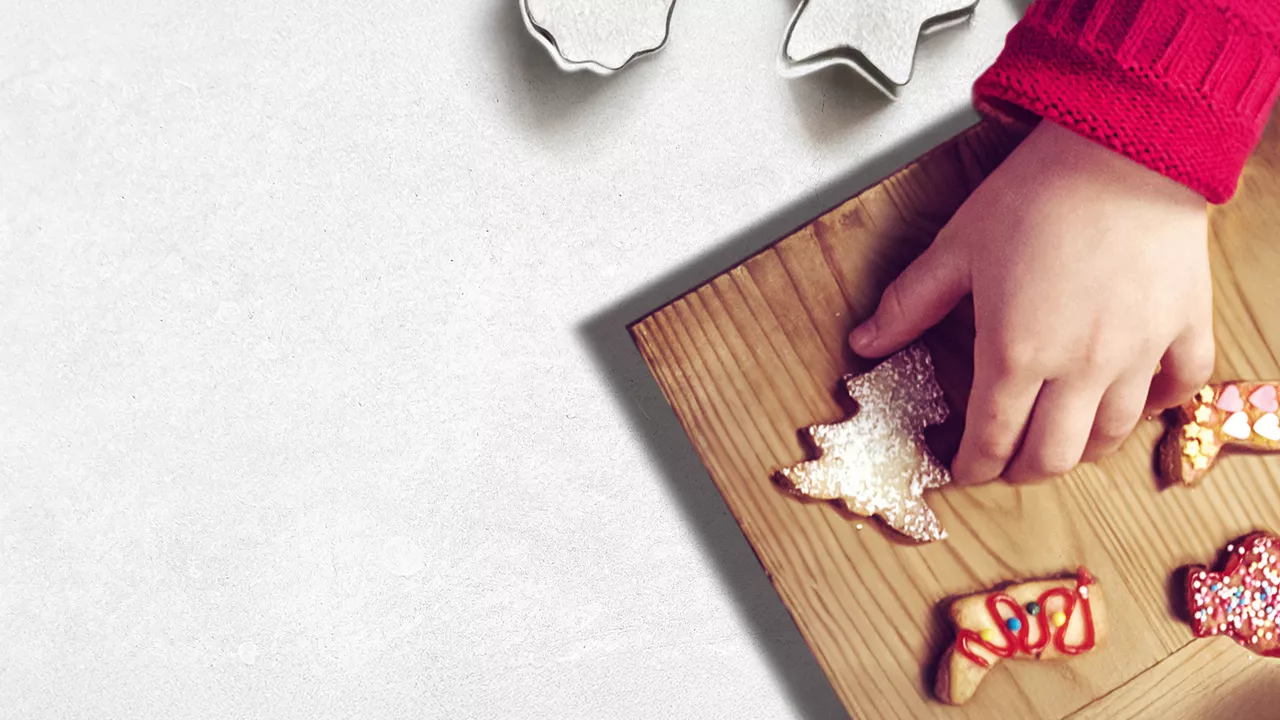 Kind greift nach einem Tannenbaum-Plätzchen auf einem Holzbrett, umgeben von Ausstechformen und verzierten Keksen.