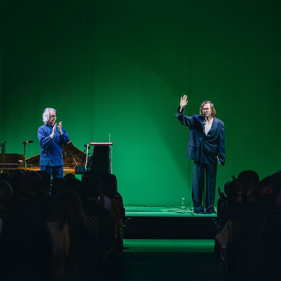 Zwei Männer stehen auf einer Bühne mit grünem Hintergrund; einer winkt, der andere applaudiert, Publikum im Vordergrund.