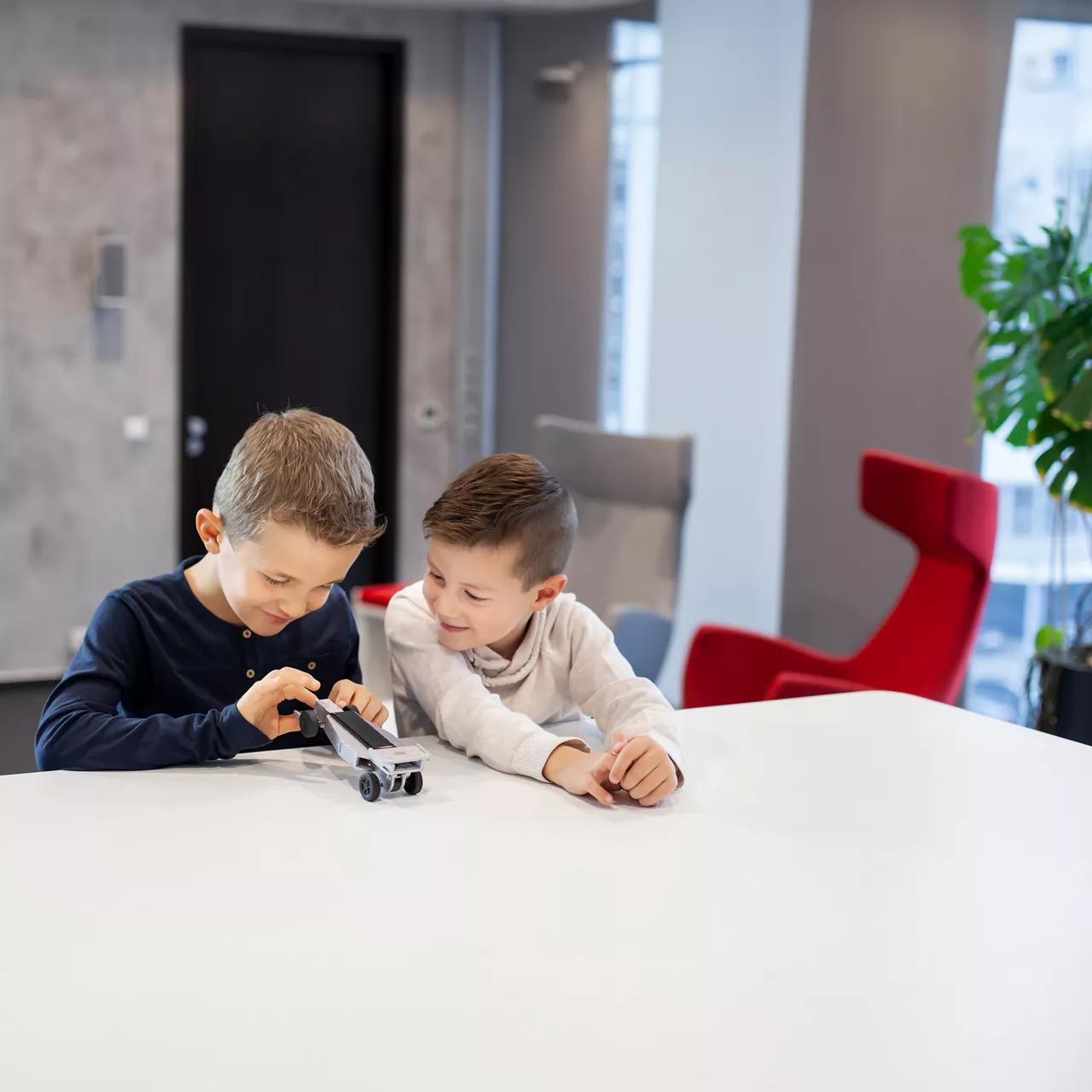 Zwei Jungen spielen lächelnd mit einem kleinen Modellauto auf einem weißen Tisch in modernem Innenraum.