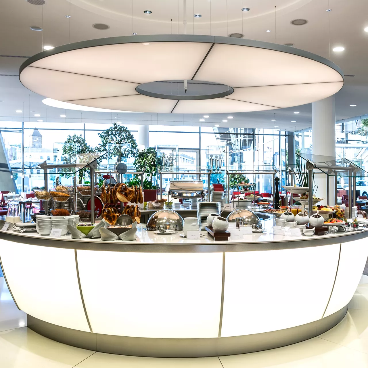 Modernes Buffet mit Tellern, Brezeln, Obst und Speisen in hellem Restaurant mit großen Fenstern und runder Deckenbeleuchtung.