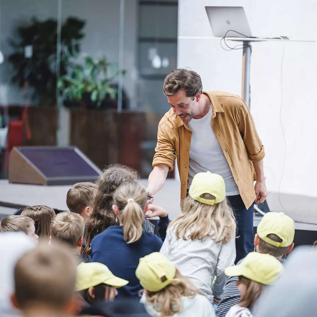 Ein Mann im braunen Hemd beugt sich vor und begrüßt eine Gruppe von Kindern, die auf einem Event versammelt sind.
