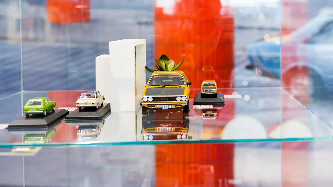 Verschiedene Modellautos, darunter ein gelber Audi 80 GTE, in einer Glasvitrine mit rotem Hintergrund ausgestellt.