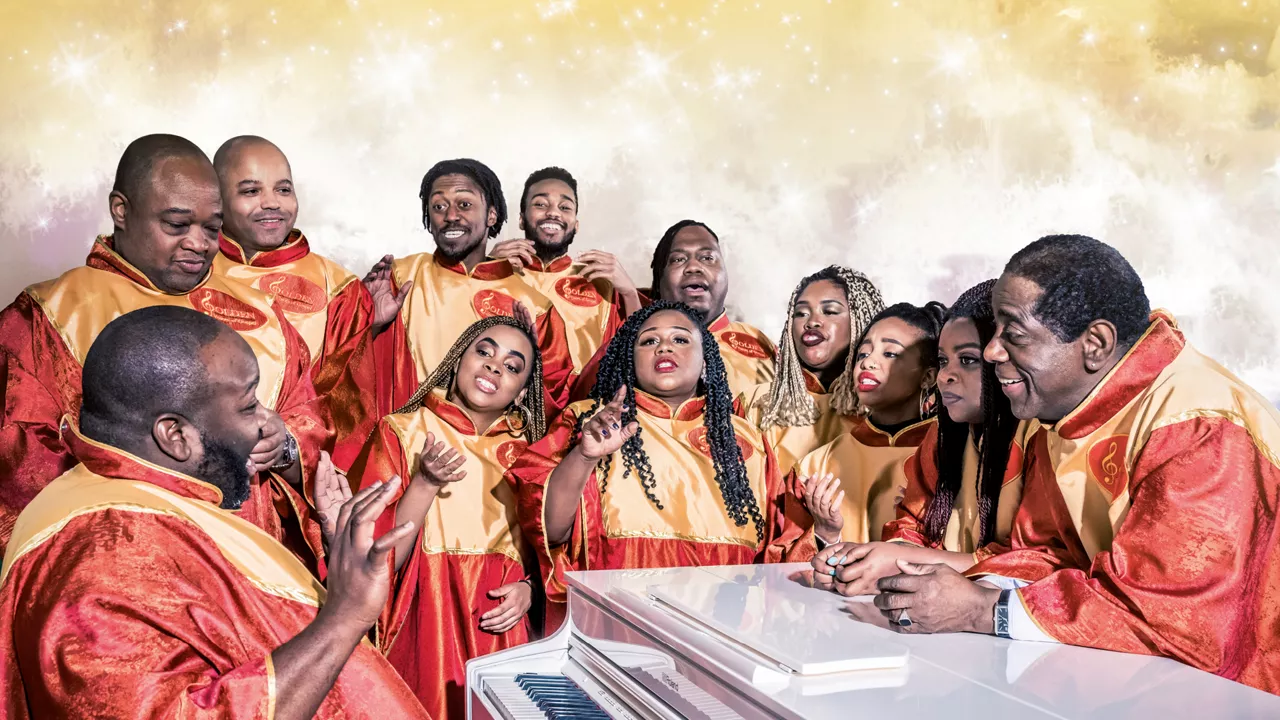 Gruppe von Sängerinnen und Sängern in roten und goldenen Chorgewändern sitzt singend um ein weißes Klavier vor festlichem Hintergrund.