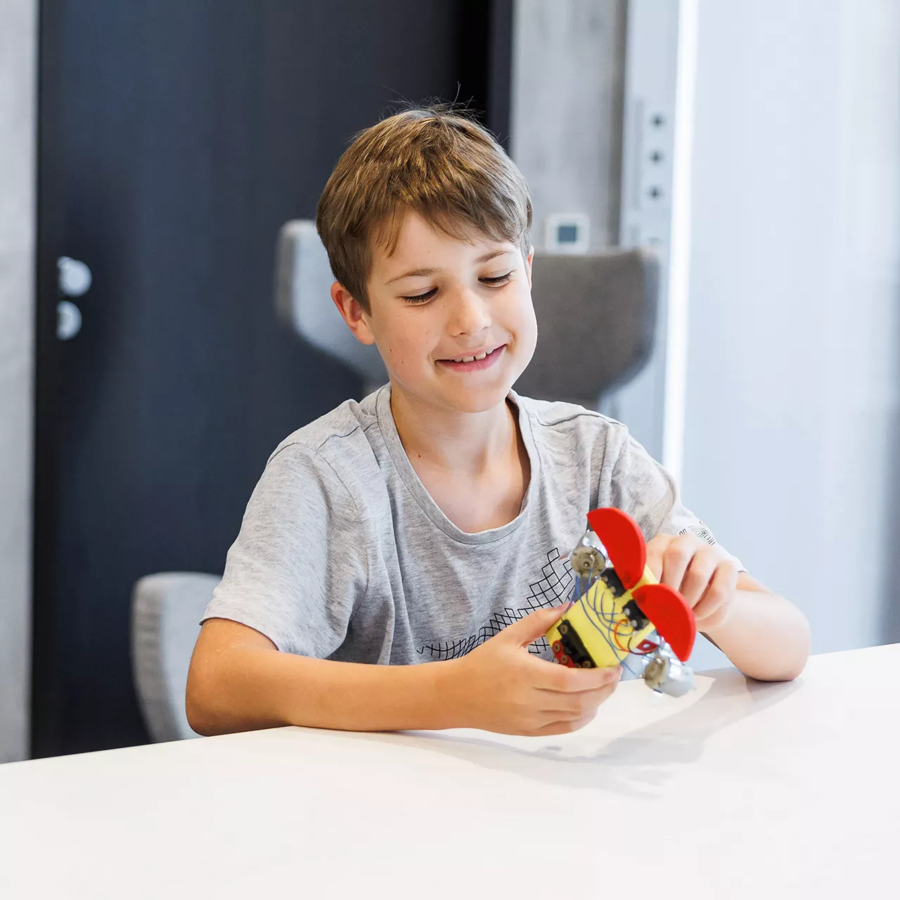 Junge sitzt an einem Tisch und hält einen zusammengebauten Gegenstand in den Händen und lächelt