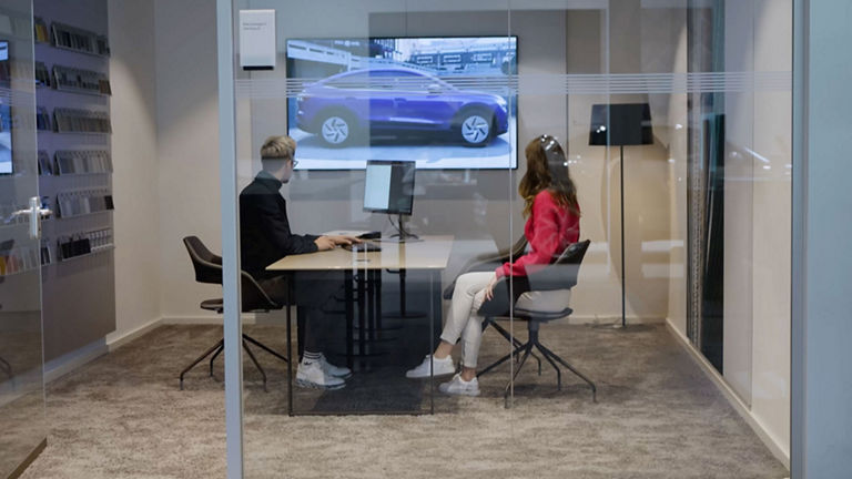 Zwei Menschen sitzen sich an einem Schreibtisch gegenüber und betrachten auf einem second Screen einen Audi.