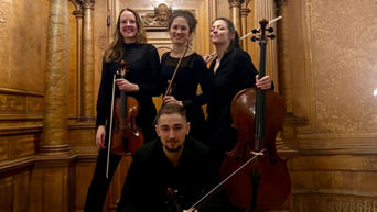 Aeterna Quartett posiert vor einer hölzernen Wand mit Instrumenten in den Häden (3 Frauen hinten stehend und ein Mann, vorne sitzen, in schwarzen Outfits)