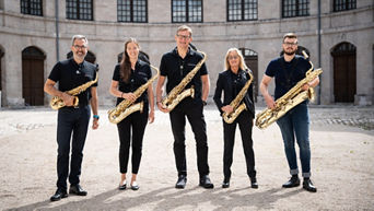 Das Saxophon-Ensemble steht mit Saxophonen und schwarzen Outfit vor einem alten, leer stehenden Gebäude (beige Mauern)