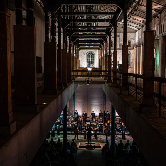 Chor und Dirigent in einer dramatisch beleuchteten Industriehalle mit hohen Säulen und Publikum.
