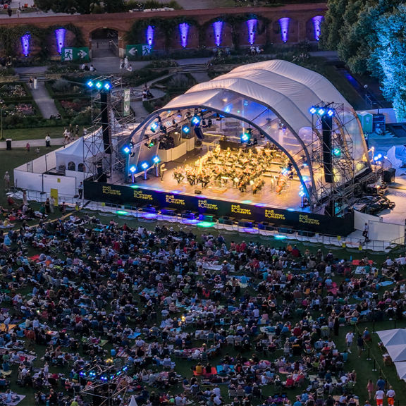 Open-Air-Konzert bei Nacht mit großer Bühne, bunten Lichtern und vielen Zuschauern auf einer Wiese.