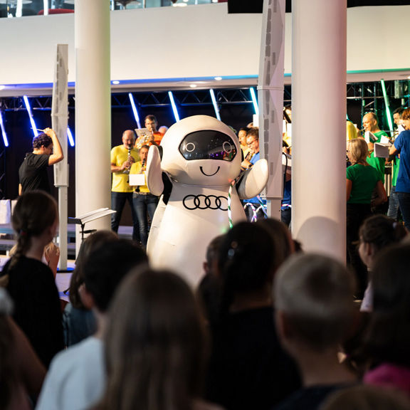 Kinder stehen vor einer Bühne mit einer Roboterfigur und Musikern bei einem Event der Audi Sommerkonzerte.