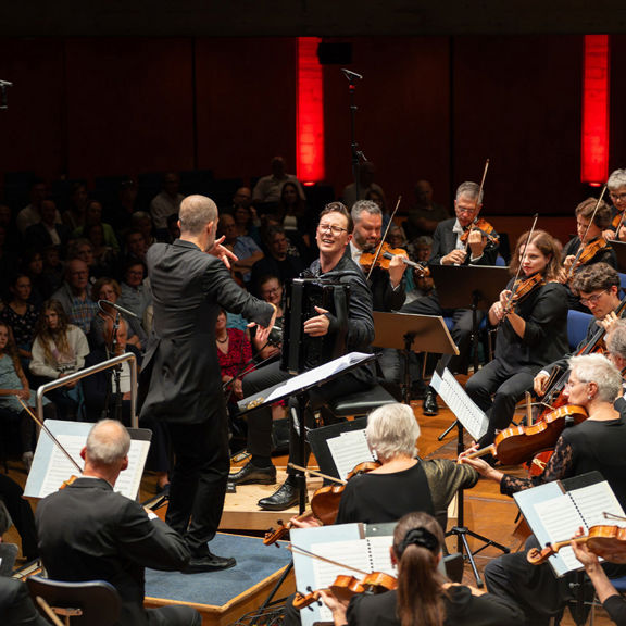 Orchester mit Dirigent und Akkordeonspieler auf Bühne vor großem Publikum in einem Konzertsaal.