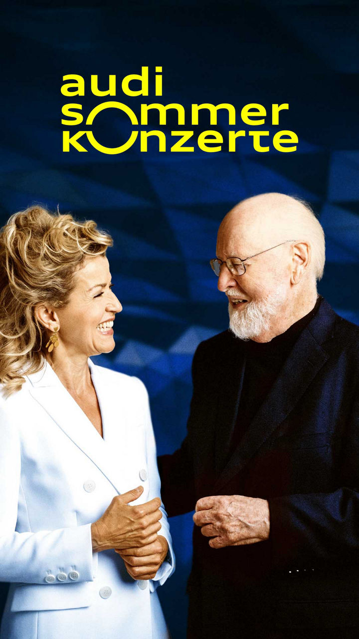 Ältere Dame und älterer Herr schauen sich an und lächeln, im Hintergrund blau und das gelbe Logo der Audi Sommerkonzerte