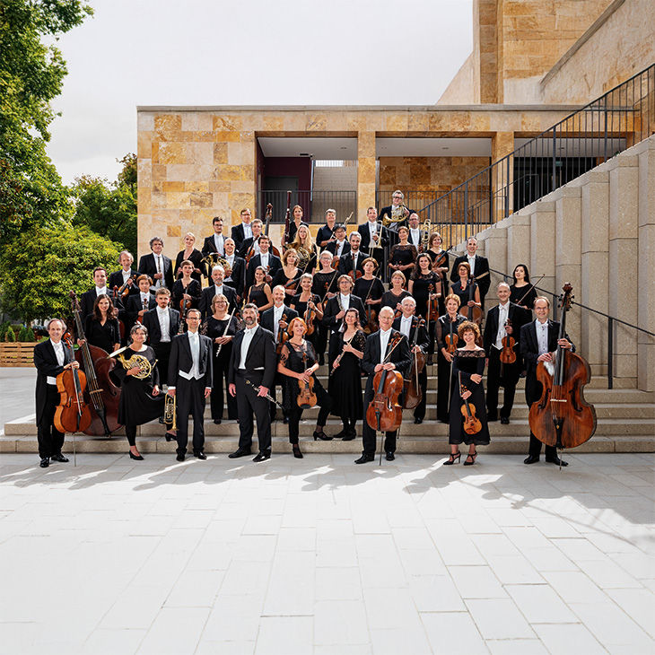 Eine große Gruppe von Musikerinnen und Musikern mit Instrumenten posiert in eleganter Kleidung vor einem modernen Gebäude.