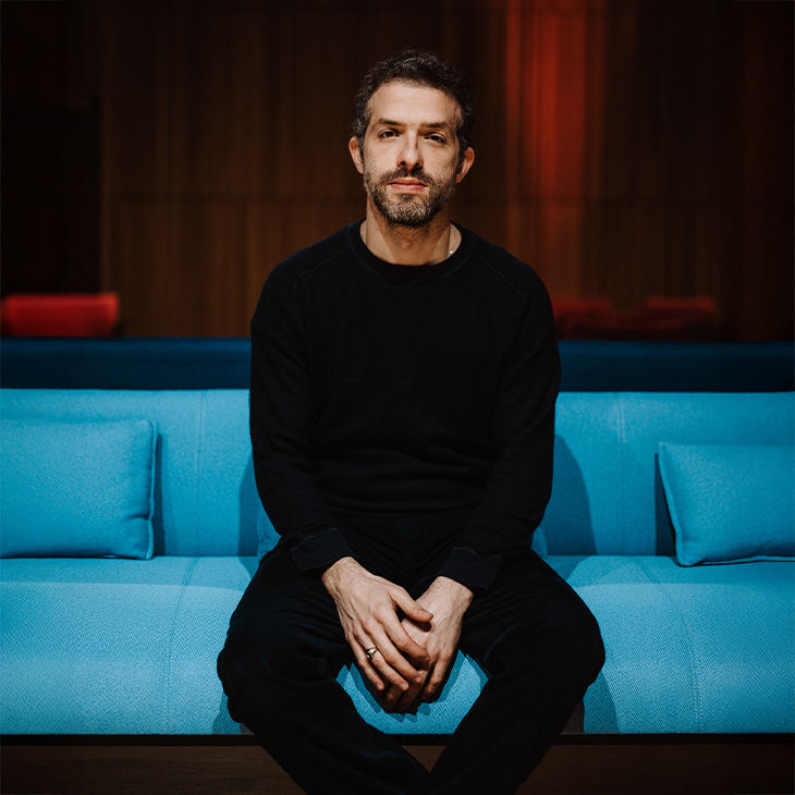 Ein Mann in schwarzer Kleidung sitzt auf einem blauen Sofa und blickt ruhig in die Kamera.
