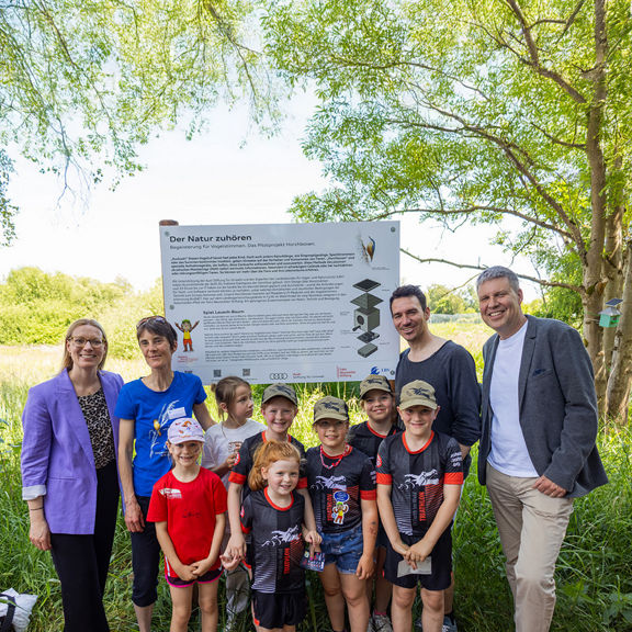 Erwachsene und Kinder posieren gemeinsam vor einer Infotafel zum Projekt „Horchboxen“ auf einer Wiese.