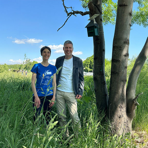 Zwei Erwachsene stehen lächelnd neben einer grün-schwarzen Horchbox mit Solarpanel, die an einem Baum befestigt ist.