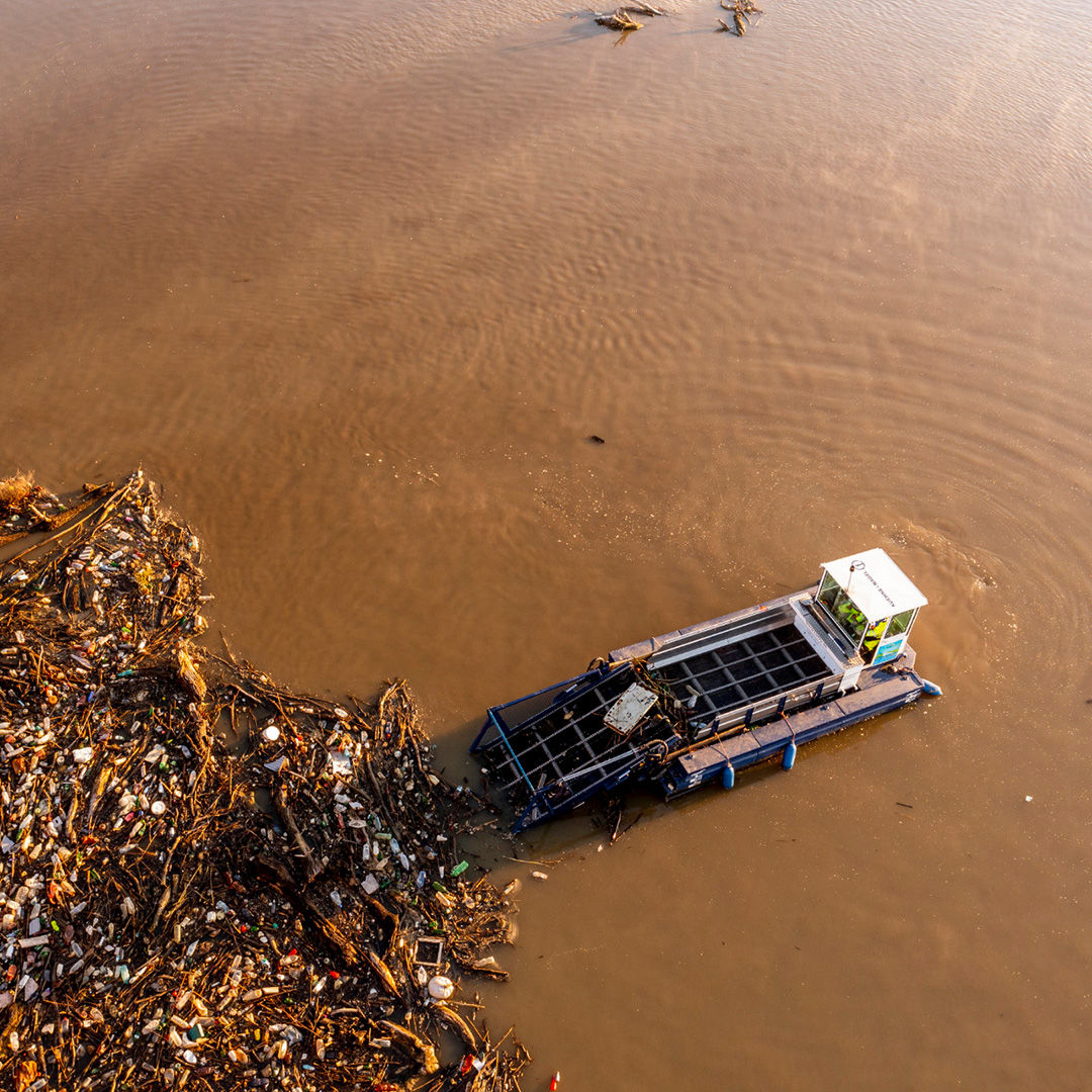 Vogelperspektive auf einen Fluss mit braun gefärbten Wasser. Ein Müllsammelboot nimmt Unrat aus dem Wasser auf.