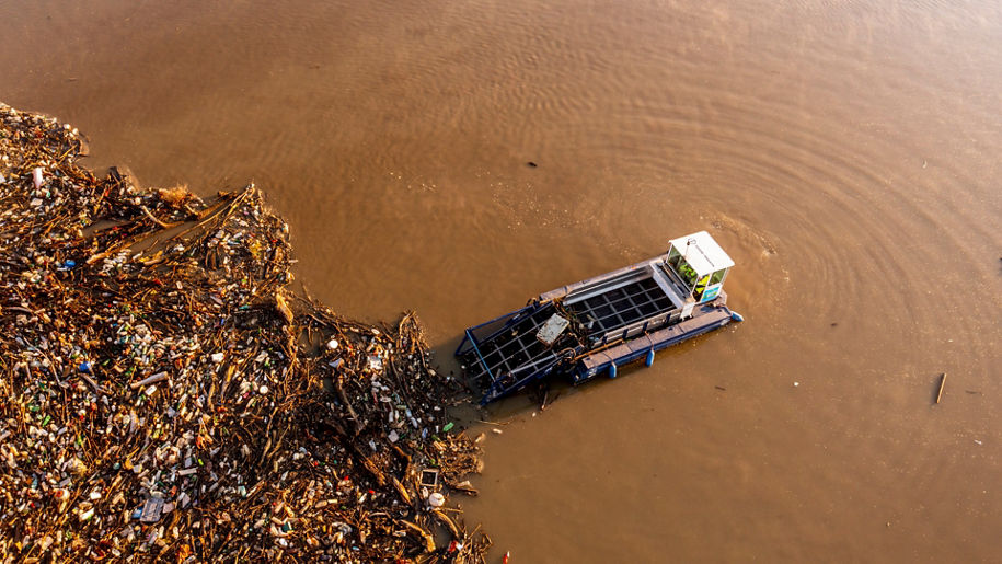 Vogelperspektive auf einen Fluss mit braun gefärbten Wasser. Ein Müllsammelboot nimmt Unrat aus dem Wasser auf.