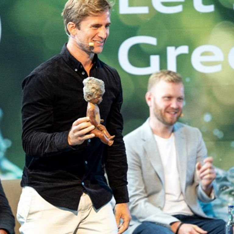 Mann hält auf einer Bühne ein Puppe in der Hand. Im Hintergrund ist der Greenovation Schriftzug zu sehen.