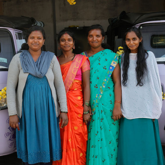Four women stand side by side in front of decorated lilac electric rickshaws, smiling at the camera.